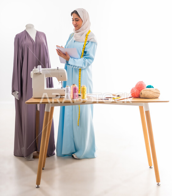 اختراع وابتكار تصاميم جديدة وعصرية، بورتريه لمصممة عربية خليجية عمانية محجبة ترتدي عباءة زرقاء تقوم برسم تصميمات معينة، أهمية المرأة في مجال الخياطة، ورشة عمل تصميم ازياء، مهن وأعمال فنية نسائية، فنون ومهارات تفصيل الملابس النسائية، مفهوم عالم الأزياء والموضة