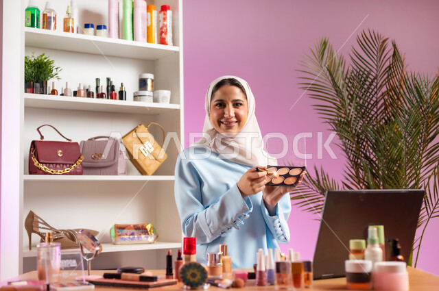 مكياج وميك اب تتوريال أون لاين, مفهوم الانوثه والاهتمام بالمظهر الخارجي, إمرأة عربية خليجية عمانية ترتدي عباءة زرقاء تجلس أمام الحاسوب وتحمل بيدها أحد معدات الميكاب، أدوات تجميل نسائيه عصريه، التسويق لمنتجات التجميل عبر وسائل التواصل الاجتماعي, صالون نسائي راقي