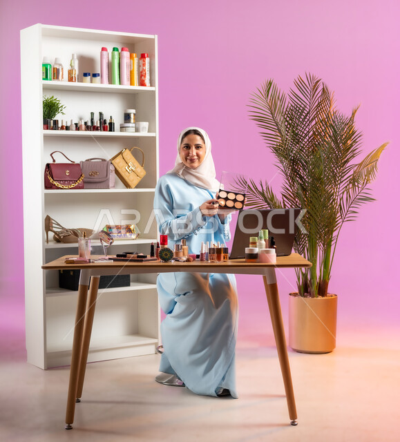 مكياج وميك اب تتوريال أون لاين, مفهوم الانوثه والاهتمام بالمظهر الخارجي, إمرأة عربية خليجية عمانية ترتدي عباءة زرقاء تجلس أمام الحاسوب وتحمل بيدها أحد معدات الميكاب، أدوات تجميل نسائيه عصريه، التسويق لمنتجات التجميل عبر وسائل التواصل الاجتماعي, صالون نسائي راقي