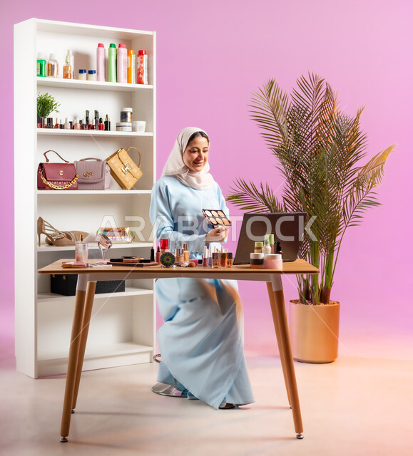 مكياج وميك اب تتوريال أون لاين, مفهوم الانوثه والاهتمام بالمظهر الخارجي, إمرأة عربية خليجية عمانية ترتدي عباءة زرقاء تجلس أمام الحاسوب وتحمل بيدها أحد معدات الميكاب، أدوات تجميل نسائيه عصريه، التسويق لمنتجات التجميل عبر وسائل التواصل الاجتماعي, صالون نسائي راقي