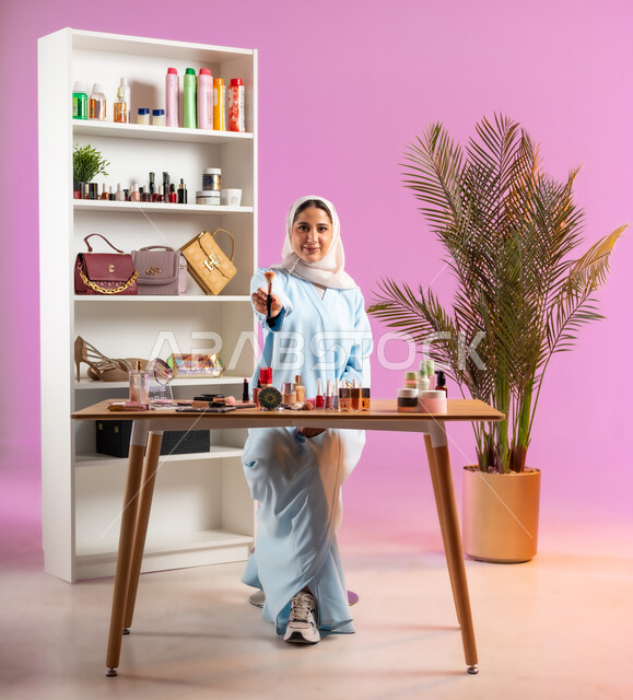 أدوات تجميل نسائيه عصريه، إمرأة عربية خليجية عمانية ترتدي عباءة زرقاء تنظر إلى الكاميرا بايماءات الابتسامه تشير بيدها لأحد أدوات الميك اب،مفهوم الانوثه والاهتمام بالمظهر الخارجي, التسويق لمنتجات التجميل, مكياج وميكاب تتوريال, صالون نسائي راقي