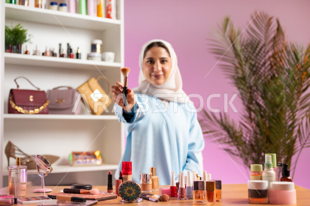 أدوات تجميل نسائيه عصريه، إمرأة عربية خليجية عمانية ترتدي عباءة زرقاء تنظر إلى الكاميرا بايماءات الابتسامه تشير بيدها لأحد أدوات الميك اب،مفهوم الانوثه والاهتمام بالمظهر الخارجي, التسويق لمنتجات التجميل, مكياج وميكاب تتوريال, صالون نسائي راقي