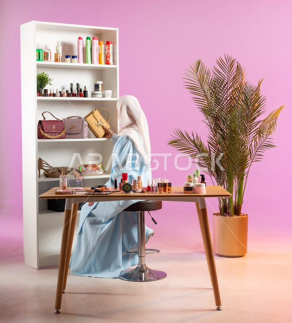 ترتيب وتنسيق الرفوف، صورة من الخلف لإمرأة عربية خليجية عمانية محجبة ترتدي عباءة زرقاء تجلس على كرسي أمام خزانة للاكسسوارات، مفهوم الانوثه والاهتمام بالمظهر الخارجي, أدوات تجميل نسائيه عصريه، التسويق لمنتجات التجميل, مكياج وميكاب تتوريال, صالون نسائي راقي