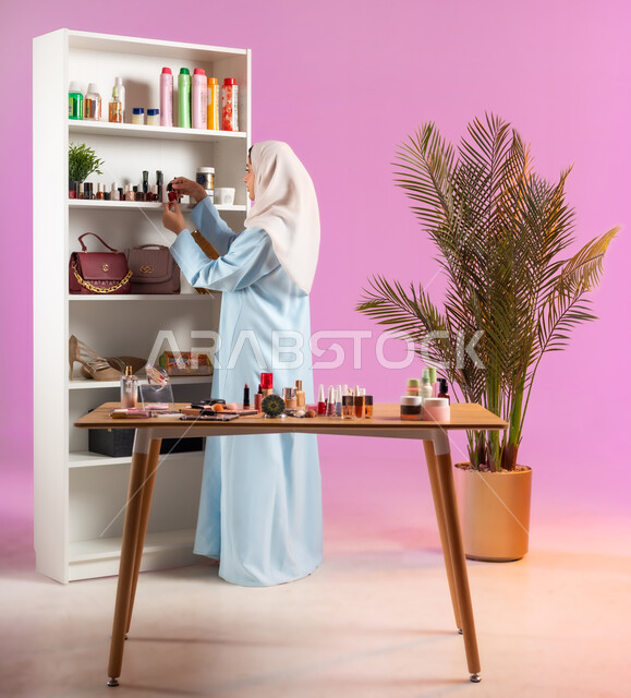 رف يحتوي على ألوان مختلفة من طلاء الأظافر، مرأة عربية خليجية عمانية محجبة ترتدي عباءة زرقاء تقوم باختيار لون مناكير مناسب،مفهوم الانوثه والاهتمام بالمظهر الخارجي, أدوات تجميل نسائيه عصريه، التسويق لمنتجات التجميل, مكياج وميكاب تتوريال, صالون نسائي راقي