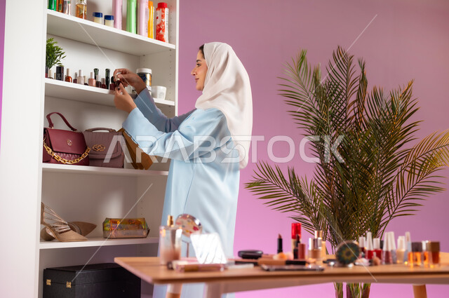 رف يحتوي على ألوان مختلفة من طلاء الأظافر، مرأة عربية خليجية عمانية محجبة ترتدي عباءة زرقاء تقوم باختيار لون مناكير مناسب،مفهوم الانوثه والاهتمام بالمظهر الخارجي, أدوات تجميل نسائيه عصريه، التسويق لمنتجات التجميل, مكياج وميكاب تتوريال, صالون نسائي راقي