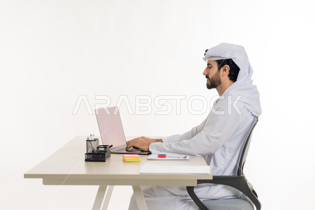أداء المهام والاعمال المكتبيه داخل مقر الشركه، بورتريه لرجل عربي خليجي اماراتي يرتدي الكندوره والغتره البيضاء يجلس خلف مكتبه ويقوم بالعمل على جهاز الاب توب المحمول، دراسه المشاريع ومراجعه التقارير السنويه، الاشراف على المخططات وتحليل اداء العمل، جلسه عمل عن بعد، خلفيه بيضاء