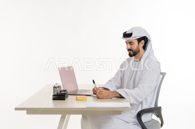 أداء المهام والاعمال المكتبيه داخل مقر الشركه، بورتريه لرجل عربي خليجي اماراتي يرتدي الكندوره والغتره البيضاء يجلس خلف مكتبه ويقوم بالعمل على جهاز الاب توب المحمول، دراسه المشاريع ومراجعه التقارير السنويه، الاشراف على المخططات وتحليل اداء العمل، جلسه عمل عن بعد، خلفيه بيضاء