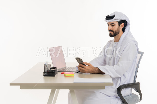 أداء المهام والاعمال المكتبيه داخل مقر الشركه، بورتريه لرجل عربي خليجي اماراتي يرتدي الكندوره والغتره البيضاء يجلس خلف مكتبه ويقوم بالعمل على جهاز الاب توب المحمول، دراسه المشاريع ومراجعه التقارير السنويه، الاشراف على المخططات وتحليل اداء العمل، جلسه عمل عن بعد، خلفيه بيضاء
