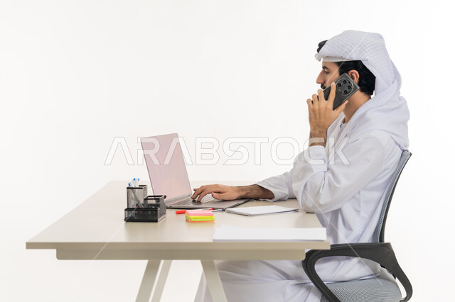 أداء المهام والاعمال المكتبيه داخل مقر الشركه، بورتريه لرجل عربي خليجي اماراتي يرتدي الكندوره والغتره البيضاء يجلس خلف مكتبه ويقوم بالعمل على جهاز الاب توب المحمول، دراسه المشاريع ومراجعه التقارير السنويه، الاشراف على المخططات وتحليل اداء العمل، جلسه عمل عن بعد، خلفيه بيضاء