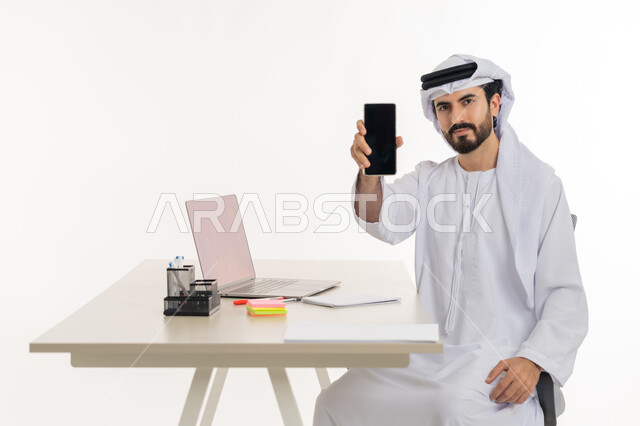 Performing office tasks and work inside the company headquarters, portrait of an Arab Gulf Emirati man wearing a white kandura and ghutra sitting behind his desk and working on a laptop, studying projects and reviewing annual reports, supervising plans and analyzing work performance, remote work session, white background