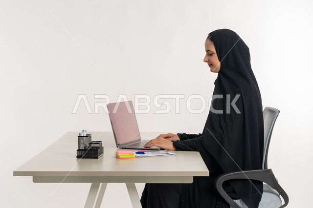 أداء المهام والاعمال المكتبيه داخل مقر الشركه، بورتريه لسيده عربيه خليجيه اماراتيه ترتدي العباءه والحجاب الاماراتي تجلس خلف المكتب وتقوم بالعمل على جهاز الاب توب المحمول، دراسه المشاريع ومراجعه التقارير السنويه، الاشراف على المخططات وتحليل اداء العمل، جلسه عمل عن بعد، خلفيه بيضاء