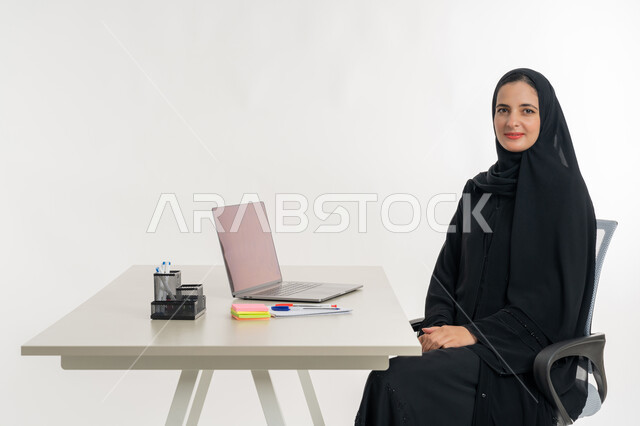 Performing tasks and office work inside the company's headquarters, portrait of an Arab Gulf Emirati woman wearing an abaya and Emirati hijab sitting behind a desk and working on a laptop, studying projects and reviewing annual reports, supervising plans and analyzing work performance, remote work session, white background