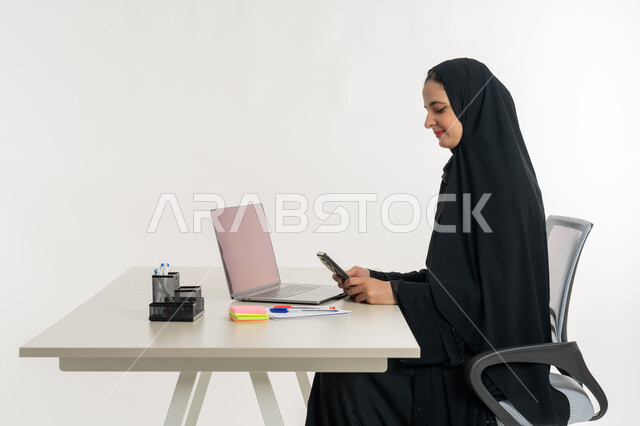 أداء المهام والاعمال المكتبيه داخل مقر الشركه، بورتريه لسيده عربيه خليجيه اماراتيه ترتدي العباءه والحجاب الاماراتي تجلس خلف المكتب وتقوم بالعمل على جهاز الاب توب المحمول، دراسه المشاريع ومراجعه التقارير السنويه، الاشراف على المخططات وتحليل اداء العمل، جلسه عمل عن بعد، خلفيه بيضاء