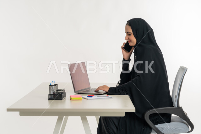 أداء المهام والاعمال المكتبيه داخل مقر الشركه، بورتريه لسيده عربيه خليجيه اماراتيه ترتدي العباءه والحجاب الاماراتي تجلس خلف المكتب وتقوم بالعمل على جهاز الاب توب المحمول، دراسه المشاريع ومراجعه التقارير السنويه، الاشراف على المخططات وتحليل اداء العمل، جلسه عمل عن بعد، خلفيه بيضاء