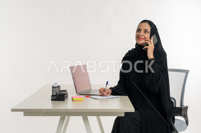 أداء المهام والاعمال المكتبيه داخل مقر الشركه، بورتريه لسيده عربيه خليجيه اماراتيه ترتدي العباءه والحجاب الاماراتي تجلس خلف المكتب وتقوم بالعمل على جهاز الاب توب المحمول، دراسه المشاريع ومراجعه التقارير السنويه، الاشراف على المخططات وتحليل اداء العمل، جلسه عمل عن بعد، خلفيه بيضاء
