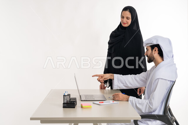مهن ووظائف مكتبية والعمل عن بعد، بورتريه لرجل عربي خليجي اماراتي يرتدي الكندوره والغتره البيضاء الاماراتيه يجلس خلف مكتبه ويقوم بالعمل على جهاز الاب توب المحمول بمساعده زميلته لدراسه وتحليل المشاريع الجديده ومناقشه اداء وتقارير العمل، خلفيه بيضاء