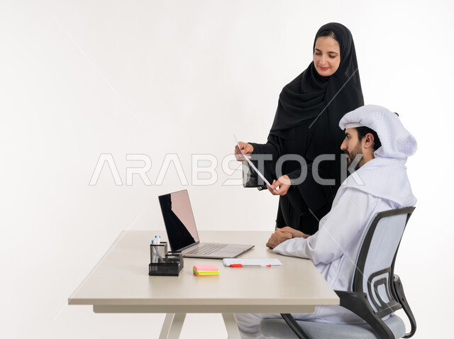 مهن ووظائف مكتبية والعمل عن بعد، بورتريه لرجل عربي خليجي اماراتي يرتدي الكندوره والغتره البيضاء الاماراتيه يجلس خلف مكتبه ويقوم بالعمل على جهاز الاب توب المحمول بمساعده زميلته لدراسه وتحليل المشاريع الجديده ومناقشه اداء وتقارير العمل، خلفيه بيضاء