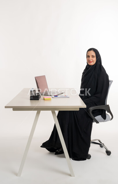 أداء المهام والاعمال المكتبيه داخل مقر الشركه، بورتريه مقرب لسيده عربيه خليجيه اماراتيه ترتدي العباءه والحجاب الاماراتي تجلس خلف المكتب وتقوم بالعمل على جهاز الاب توب المحمول، دراسه المشاريع ومراجعه التقارير السنويه، الاشراف على المخططات وتحليل اداء العمل، جلسه عمل عن بعد، خلفيه بيضاء
