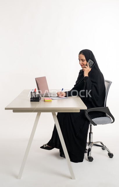 Performing tasks and office work inside the company headquarters, close-up portrait of an Arab Gulf Emirati woman wearing an abaya and Emirati hijab sitting behind a desk and working on a laptop, studying projects and reviewing annual reports, supervising plans and analyzing work performance, remote work session, white background