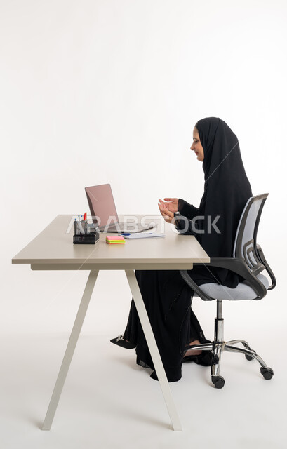 أداء المهام والاعمال المكتبيه داخل مقر الشركه، بورتريه مقرب لسيده عربيه خليجيه اماراتيه ترتدي العباءه والحجاب الاماراتي تجلس خلف المكتب وتقوم بالعمل على جهاز الاب توب المحمول، دراسه المشاريع ومراجعه التقارير السنويه، الاشراف على المخططات وتحليل اداء العمل، جلسه عمل عن بعد، خلفيه بيضاء