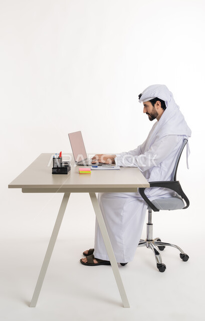 أداء المهام والاعمال المكتبيه داخل مقر الشركه، بورتريه مقرب لرجل عربي خليجي اماراتي يرتدي الكندوره والغتره البيضاء الاماراتيه يجلس خلف المكتب ويقوم بالعمل على جهاز الاب توب المحمول، دراسه المشاريع ومراجعه التقارير السنويه، الاشراف على المخططات وتحليل اداء العمل، جلسه عمل عن بعد، خلفيه بيضاء