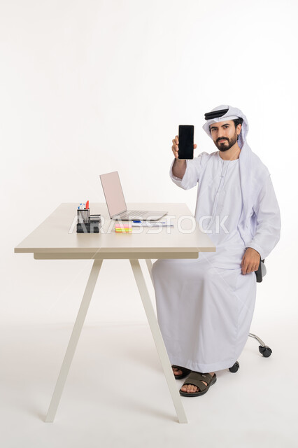 أداء المهام والاعمال المكتبيه داخل مقر الشركه، بورتريه مقرب لرجل عربي خليجي اماراتي يرتدي الكندوره والغتره البيضاء الاماراتيه يجلس خلف المكتب ويقوم بالعمل على جهاز الاب توب المحمول، دراسه المشاريع ومراجعه التقارير السنويه، الاشراف على المخططات وتحليل اداء العمل، جلسه عمل عن بعد، خلفيه بيضاء