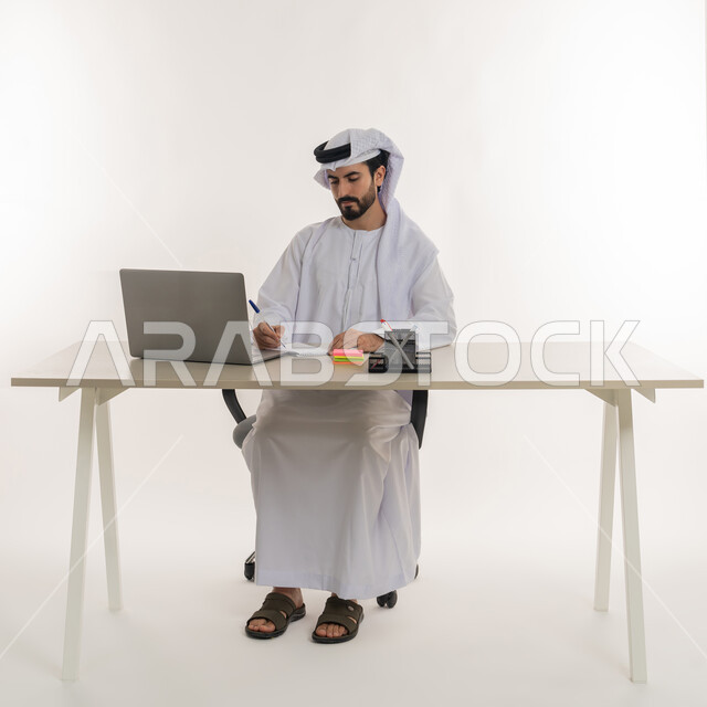 أداء المهام والاعمال المكتبيه داخل مقر الشركه، بورتريه مقرب لرجل عربي خليجي اماراتي يرتدي الكندوره والغتره البيضاء الاماراتيه يجلس خلف المكتب ويقوم بالعمل على جهاز الاب توب المحمول، دراسه المشاريع ومراجعه التقارير السنويه، الاشراف على المخططات وتحليل اداء العمل، جلسه عمل عن بعد، خلفيه بيضاء