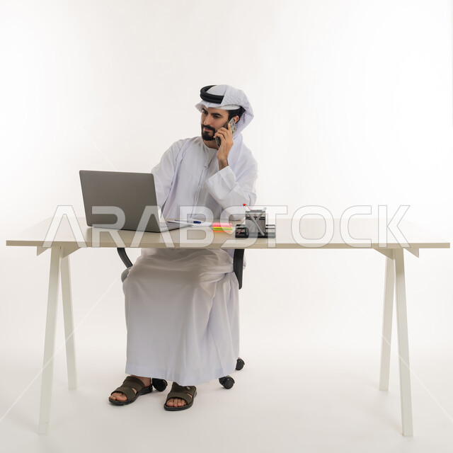 أداء المهام والاعمال المكتبيه داخل مقر الشركه، بورتريه مقرب لرجل عربي خليجي اماراتي يرتدي الكندوره والغتره البيضاء الاماراتيه يجلس خلف المكتب ويقوم بالعمل على جهاز الاب توب المحمول، دراسه المشاريع ومراجعه التقارير السنويه، الاشراف على المخططات وتحليل اداء العمل، جلسه عمل عن بعد، خلفيه بيضاء