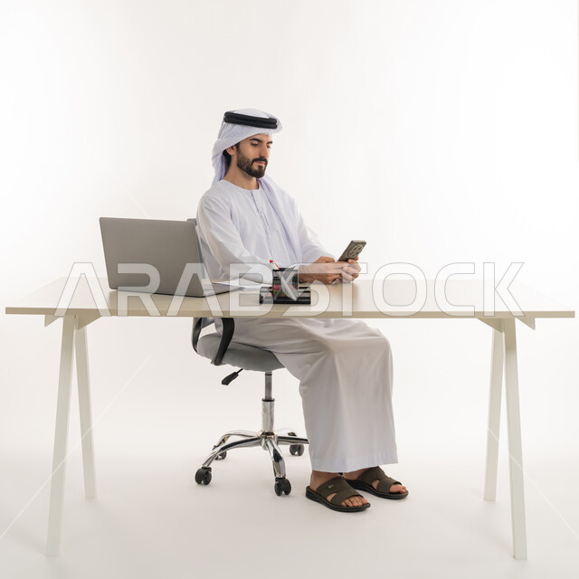 أداء المهام والاعمال المكتبيه داخل مقر الشركه، بورتريه مقرب لرجل عربي خليجي اماراتي يرتدي الكندوره والغتره البيضاء الاماراتيه يجلس خلف المكتب ويقوم بالعمل على جهاز الاب توب المحمول، دراسه المشاريع ومراجعه التقارير السنويه، الاشراف على المخططات وتحليل اداء العمل، جلسه عمل عن بعد، خلفيه بيضاء