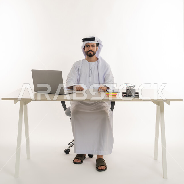 أداء المهام والاعمال المكتبيه داخل مقر الشركه، بورتريه مقرب لرجل عربي خليجي اماراتي يرتدي الكندوره والغتره البيضاء الاماراتيه يجلس خلف المكتب ويقوم بالعمل على جهاز الاب توب المحمول، دراسه المشاريع ومراجعه التقارير السنويه، الاشراف على المخططات وتحليل اداء العمل، جلسه عمل عن بعد، خلفيه بيضاء