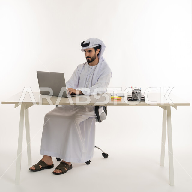 أداء المهام والاعمال المكتبيه داخل مقر الشركه، بورتريه مقرب لرجل عربي خليجي اماراتي يرتدي الكندوره والغتره البيضاء الاماراتيه يجلس خلف المكتب ويقوم بالعمل على جهاز الاب توب المحمول، دراسه المشاريع ومراجعه التقارير السنويه، الاشراف على المخططات وتحليل اداء العمل، جلسه عمل عن بعد، خلفيه بيضاء