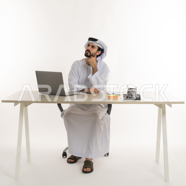 أداء المهام والاعمال المكتبيه داخل مقر الشركه، بورتريه مقرب لرجل عربي خليجي اماراتي يرتدي الكندوره والغتره البيضاء الاماراتيه يجلس خلف المكتب ويقوم بالعمل على جهاز الاب توب المحمول، دراسه المشاريع ومراجعه التقارير السنويه، الاشراف على المخططات وتحليل اداء العمل، جلسه عمل عن بعد، خلفيه بيضاء
