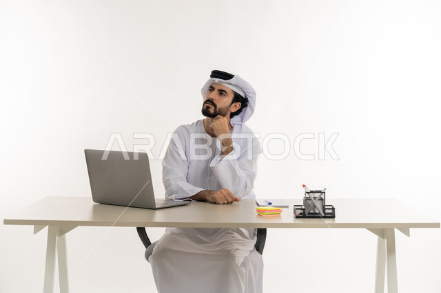 أداء المهام والاعمال المكتبيه داخل مقر الشركه، بورتريه مقرب لرجل عربي خليجي اماراتي يرتدي الكندوره والغتره البيضاء الاماراتيه يجلس خلف المكتب ويقوم بالعمل على جهاز الاب توب المحمول، دراسه المشاريع ومراجعه التقارير السنويه، الاشراف على المخططات وتحليل اداء العمل، جلسه عمل عن بعد، خلفيه بيضاء