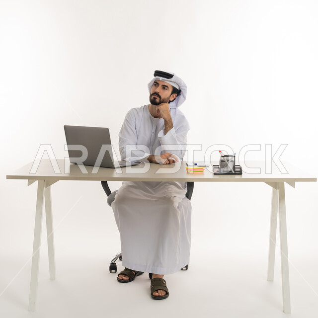 أداء المهام والاعمال المكتبيه داخل مقر الشركه، بورتريه مقرب لرجل عربي خليجي اماراتي يرتدي الكندوره والغتره البيضاء الاماراتيه يجلس خلف المكتب ويقوم بالعمل على جهاز الاب توب المحمول، دراسه المشاريع ومراجعه التقارير السنويه، الاشراف على المخططات وتحليل اداء العمل، جلسه عمل عن بعد، خلفيه بيضاء