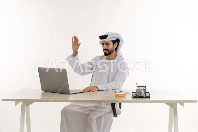 أداء المهام والاعمال المكتبيه داخل مقر الشركه، بورتريه مقرب لرجل عربي خليجي اماراتي يرتدي الكندوره والغتره البيضاء الاماراتيه يجلس خلف المكتب ويقوم بالعمل على جهاز الاب توب المحمول، دراسه المشاريع ومراجعه التقارير السنويه، الاشراف على المخططات وتحليل اداء العمل، جلسه عمل عن بعد، خلفيه بيضاء