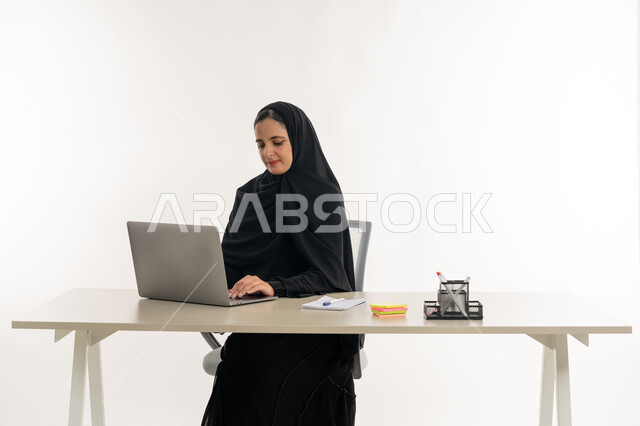 أداء المهام والاعمال المكتبيه داخل مقر الشركه، بورتريه مقرب لسيده عربيه خليجيه اماراتيه ترتدي العباءه والحجاب الاماراتي تجلس خلف المكتب وتقوم بالعمل على جهاز الاب توب المحمول، دراسه المشاريع ومراجعه التقارير السنويه، الاشراف على المخططات وتحليل اداء العمل، جلسه عمل عن بعد، خلفيه بيضاء