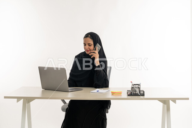 أداء المهام والاعمال المكتبيه داخل مقر الشركه، بورتريه مقرب لسيده عربيه خليجيه اماراتيه ترتدي العباءه والحجاب الاماراتي تجلس خلف المكتب وتقوم بالعمل على جهاز الاب توب المحمول، دراسه المشاريع ومراجعه التقارير السنويه، الاشراف على المخططات وتحليل اداء العمل، جلسه عمل عن بعد، خلفيه بيضاء