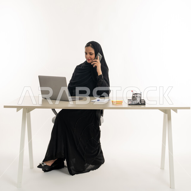 أداء المهام والاعمال المكتبيه داخل مقر الشركه، بورتريه مقرب لسيده عربيه خليجيه اماراتيه ترتدي العباءه والحجاب الاماراتي تجلس خلف المكتب وتقوم بالعمل على جهاز الاب توب المحمول، دراسه المشاريع ومراجعه التقارير السنويه، الاشراف على المخططات وتحليل اداء العمل، جلسه عمل عن بعد، خلفيه بيضاء