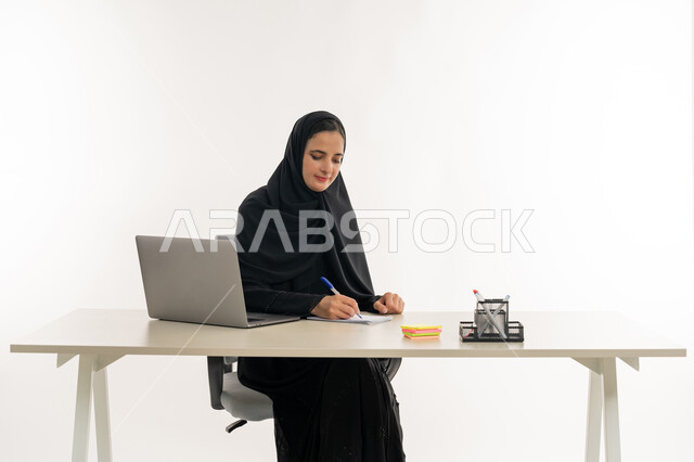 أداء المهام والاعمال المكتبيه داخل مقر الشركه، بورتريه مقرب لسيده عربيه خليجيه اماراتيه ترتدي العباءه والحجاب الاماراتي تجلس خلف المكتب وتقوم بالعمل على جهاز الاب توب المحمول، دراسه المشاريع ومراجعه التقارير السنويه، الاشراف على المخططات وتحليل اداء العمل، جلسه عمل عن بعد، خلفيه بيضاء