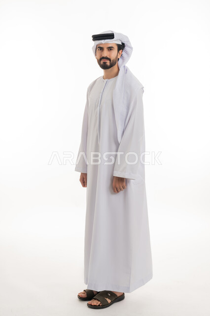 مفهوم الاناقه والرجوله والاهتمام بالمظهر الخارجي، الوقوف باستقامه والنظر الى الكاميرا بايماءات الثقه بالنفس، بورتريه بالطول الكامل لرجل عربي خليجي اماراتي يرتدي الكندوره والغتره البيضاء الاماراتيه يقف بايماءات الرجوله وعزه النفس، خلفيه بيضاء