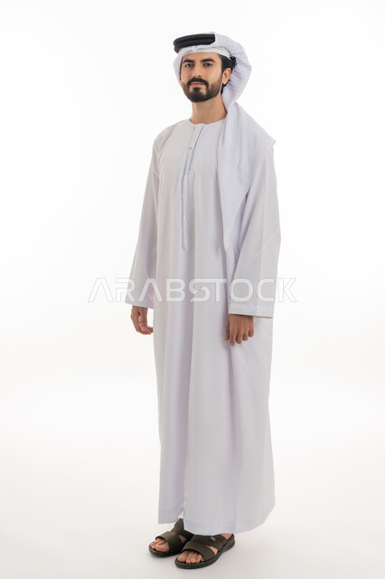 مفهوم الاناقه والرجوله والاهتمام بالمظهر الخارجي، الوقوف باستقامه والنظر الى الكاميرا بايماءات الثقه بالنفس، بورتريه بالطول الكامل لرجل عربي خليجي اماراتي يرتدي الكندوره والغتره البيضاء الاماراتيه يقف بايماءات الرجوله وعزه النفس، خلفيه بيضاء