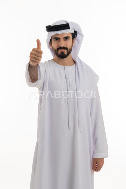 Acceptance, approval, and satisfaction with something, full length portrait of a young Arab Gulf Emirati man wearing a white kandura and ghutra pointing his thumb up with gestures of quality and excellence, white background