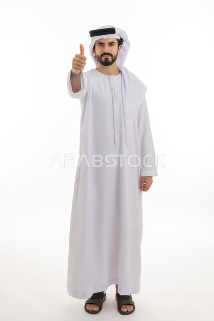 القبول والموافقه والرضا عن شئ ما, بوتريه بالطول الكامل لشاب عربي خليجي إماراتي يرتدي الكندورة والغترة البيضاء يشير باصبع الابهام نحوالاعلى بإيماءات علامه الجوده والامتياز، خلفية بيضاء