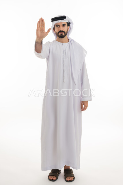 Silence and stopping doing something, standing in anger and refusing a specific order, full length portrait of a young Arab Gulf Emirati man wearing a white kandura and ghutra raising his palm up in gestures of annoyance and prohibition, requesting not to interfere and refrain from doing a specific order, white background
