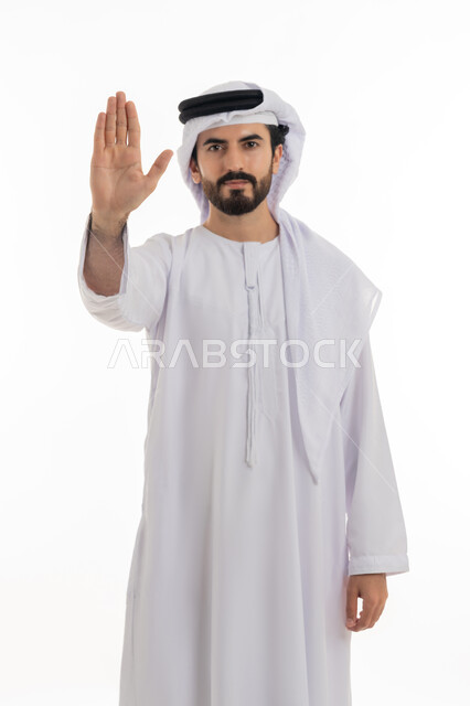 Silence and stopping doing something, standing in anger and refusing a specific order, full length portrait of a young Arab Gulf Emirati man wearing a white kandura and ghutra raising his palm up in gestures of annoyance and prohibition, requesting not to interfere and refrain from doing a specific order, white background