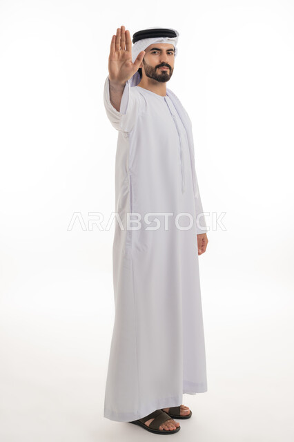 Silence and stopping doing something, standing angrily and refusing a specific order, side portrait of a young Arab Gulf Emirati man wearing a white kandura and ghutra raising his palm up with gestures of annoyance and prohibition, requesting not to interfere and refrain from doing a specific order, white background