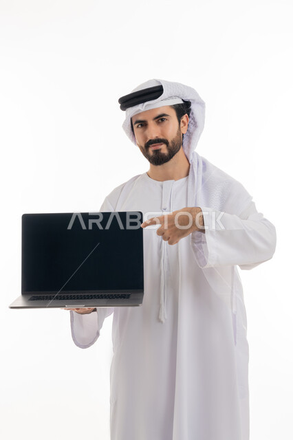 Browsing the internet and social media, using a computer to complete tasks, full-length portrait of an Emirati Gulf Arab man wearing a kandura and Emirati shemagh holding a laptop in his hand pointing at it with a blank black screen, development of technology and technical devices, white background