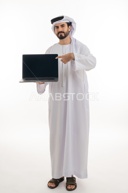 Browsing the internet and social media, using a computer to complete tasks, full-length portrait of an Emirati Gulf Arab man wearing a kandura and Emirati shemagh holding a laptop in his hand pointing at it with a blank black screen, development of technology and technical devices, white background
