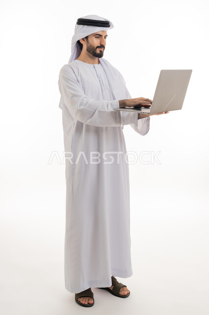 Browsing the internet and social networking programs, using the computer to complete tasks, side portrait of an Arab Gulf Emirati man wearing the Emirati kandura and shemagh, holding a laptop in his hand and working on it, development of technology and technical devices, white background