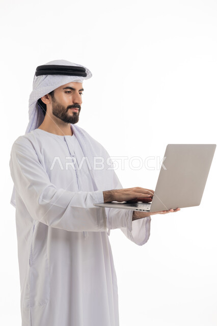 Browsing the internet and social networking programs, using the computer to complete tasks, side portrait of an Arab Gulf Emirati man wearing the Emirati kandura and shemagh, holding a laptop in his hand and working on it, development of technology and technical devices, white background